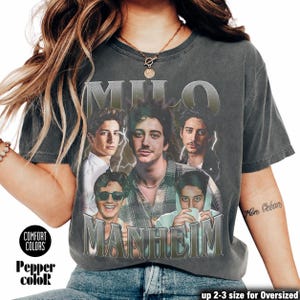 Pode incluir: Uma t-shirt de manga curta cinzento-carvão com uma impressão gráfica de Milo Manheim em várias poses e trajes. O texto MILO MANHEIM está impresso numa fonte prateada metálica acima e abaixo das imagens, com detalhes de relâmpagos.