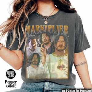 May include: Dark gray t-shirt featuring a graphic of Markiplier with the text "MARKIPLIER" in gold. The shirt has a vintage look with the words "COMFORT COLORS" and "Pepper color" printed on the bottom left.