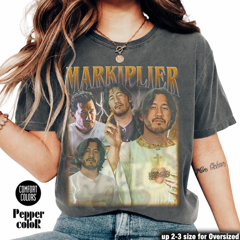 May include: Dark gray t-shirt featuring a graphic of Markiplier with the text "MARKIPLIER" in gold. The shirt has a vintage look with the words "COMFORT COLORS" and "Pepper color" printed on the bottom left.