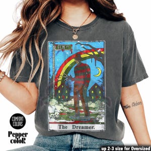 May include: Dark grey t-shirt with a horror movie graphic featuring a rainbow, houses, and the text "ELM ST." and "The Dreamer." The shirt also has the text "COMFORT COLORS" and "Pepper color."