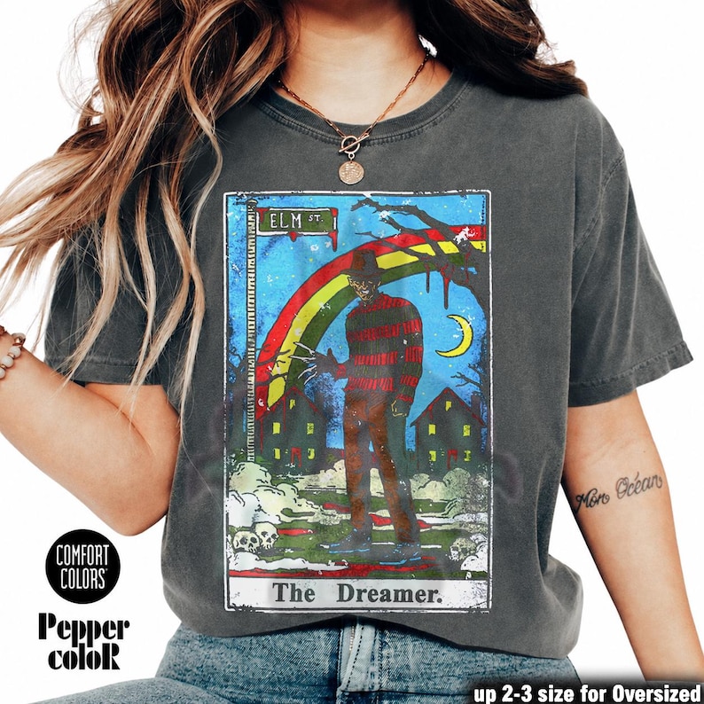 May include: Dark grey t-shirt with a horror movie graphic featuring a rainbow, houses, and the text "ELM ST." and "The Dreamer." The shirt also has the text "COMFORT COLORS" and "Pepper color."