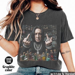 May include: A charcoal gray t-shirt featuring a graphic print of a person with dark hair wearing layered silver necklaces and rings, holding up both middle fingers. The person is surrounded by small glass bottles filled with colorful liquids.