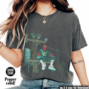 May include: A charcoal grey short-sleeve t-shirt featuring a graphic print of a red-masked character sitting on a utility pole. The graphic includes a green-tinted scene with electrical equipment and wires. The shirt has a crew neckline and a vintage wash.