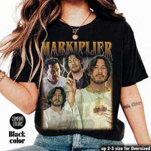 May include: Black t-shirt featuring a graphic of Markiplier in various poses with the word "MARKIPLIER" in gold lettering. The shirt has a vintage look, with the words "COMFORT COLORS" and "Black color" printed on the lower left.