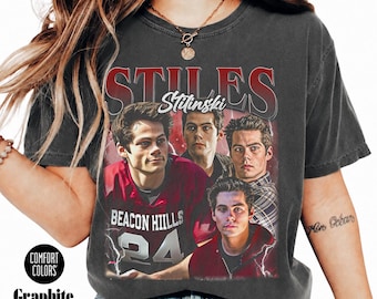 May include: A charcoal gray short-sleeve t-shirt featuring a graphic print of the character Stiles Stilinski from Teen Wolf. The design includes the text STILES and Stilinski, along with four images of the character and the text BEACON HILLS 24.