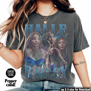 May include: Dark gray t-shirt featuring a graphic of Halle Bailey in a blue and purple design. The text "HALLE BAILEY" is printed in a blue, stylized font. The shirt has the Comfort Colors and Pepper color logos.