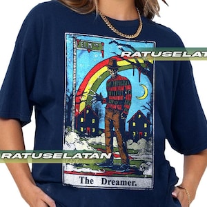 May include: Dark blue t-shirt with a horror-themed tarot card design. The image shows a figure in a striped sweater, a rainbow, houses, and the text "ELM ST" and "The Dreamer."