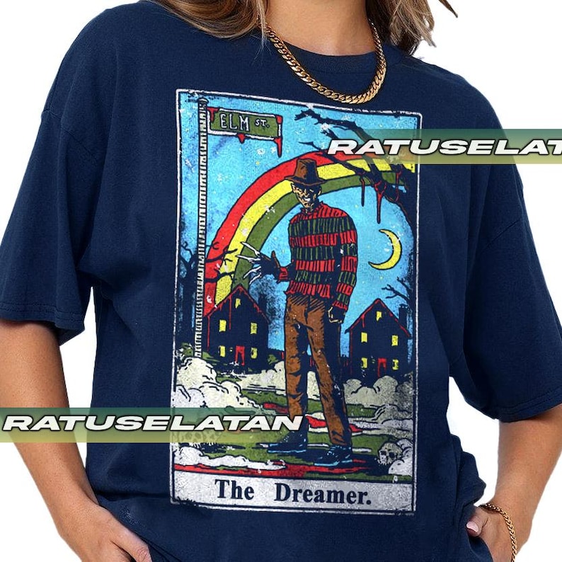 May include: Dark blue t-shirt with a horror-themed tarot card design. The image shows a figure in a striped sweater, a rainbow, houses, and the text "ELM ST" and "The Dreamer."