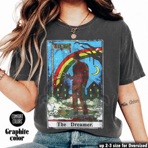 May include: Graphite-coloured t-shirt with a tarot card-style graphic. The design includes the text "ELM ST" and "The Dreamer," a rainbow, a figure with bladed fingers, and a crescent moon. The shirt is labelled "Comfort Colors."