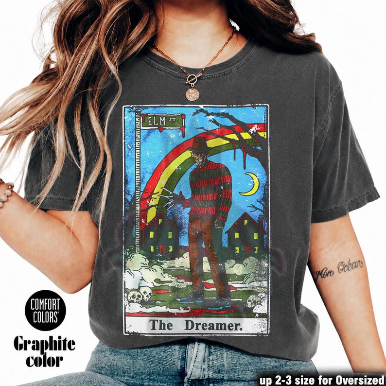 May include: Graphite-coloured t-shirt with a tarot card-style graphic. The design includes the text "ELM ST" and "The Dreamer," a rainbow, a figure with bladed fingers, and a crescent moon. The shirt is labelled "Comfort Colors."