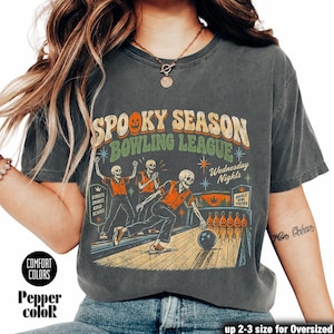 Könnte beinhalten: Ein anthrazitfarbenes T-Shirt mit einer Grafik von drei kegelnden Skeletten. Der Text lautet Spooky Season Bowling League, Wednesday Nights, Strikes Spares Good Scares und Ghouls Bowl Together. Das Design enthält orangefarbene Kürbis-Kegel.
