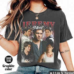 May include: A charcoal gray short-sleeve t-shirt featuring a graphic print of the character Jeremy Gilbert from The Vampire Diaries. The design includes the text JEREMY in red and Gilbert in white, surrounded by multiple images of the character.