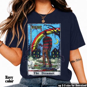 May include: Navy blue t-shirt with a tarot card design. The graphic depicts a figure in a hat and striped sweater, set against houses and a rainbow. Text includes "Elm St." and "The Dreamer".