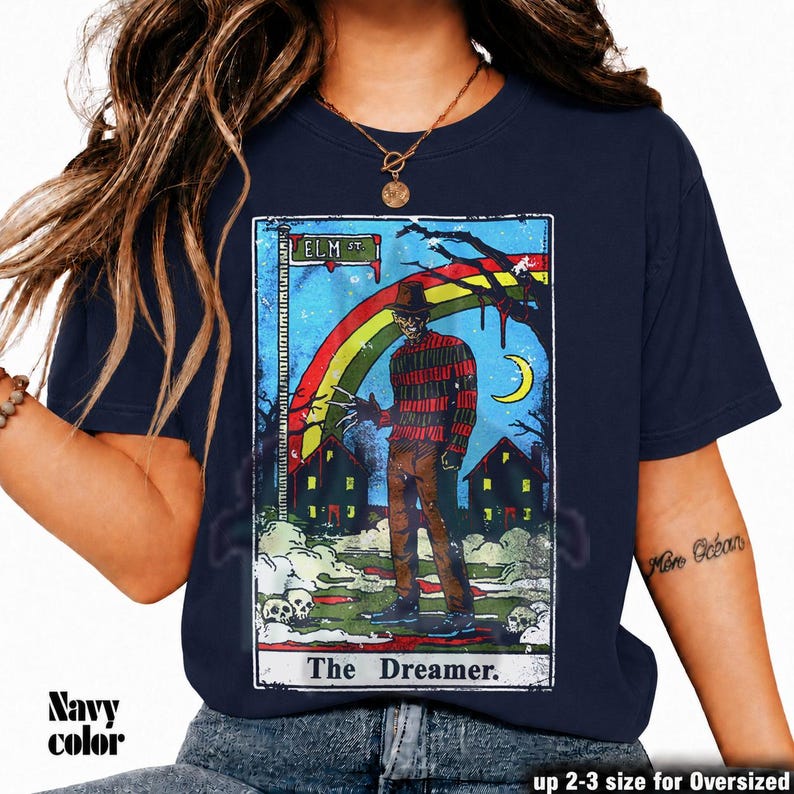 May include: Navy blue t-shirt with a tarot card design. The graphic depicts a figure in a hat and striped sweater, set against houses and a rainbow. Text includes "Elm St." and "The Dreamer".