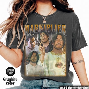 May include: Graphite-colored t-shirt featuring a vintage-style graphic of Markiplier. The design includes multiple images of Markiplier with the name "MARKIPLIER" in gold lettering. The shirt is from Comfort Colors and is available in up to 2-3 sizes for an oversized fit.