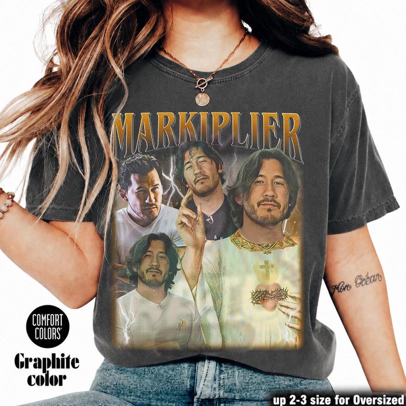 May include: Graphite-colored t-shirt featuring a vintage-style graphic of Markiplier. The design includes multiple images of Markiplier with the name "MARKIPLIER" in gold lettering. The shirt is from Comfort Colors and is available in up to 2-3 sizes for an oversized fit.