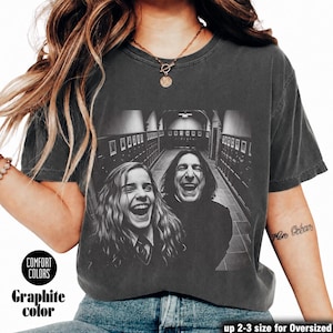 May include: A charcoal gray short-sleeve t-shirt featuring a black and white graphic print of two laughing individuals in a school hallway. The shirt has a crew neckline and a vintage-washed appearance. Text on the shirt includes Comfort Colors.