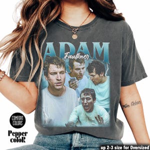 Könnte beinhalten: Dunkelgraues T-Shirt mit dem Namen "ADAM Faulkner" in hellblauer Schrift im Vintage-Stil. Das Shirt zeigt mehrere Bilder eines Mannes in verschiedenen Posen. Die Worte "COMFORT COLORS" und "Pepper coloR" sind aufgedruckt.
