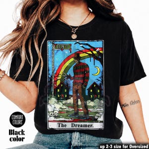 May include: Black t-shirt with a graphic of a figure in a striped sweater and hat, holding bladed gloves, with a rainbow and houses. Text reads "ELM ST" and "The Dreamer." The shirt is labelled "Comfort Colors" and "Black color."