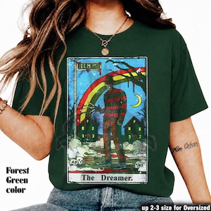 May include: Forest green t-shirt with a tarot card design. The graphic features the text "ELM ST" and "The Dreamer," along with a figure in a striped sweater, a rainbow, and a crescent moon. Available in up to 2-3 sizes for an oversized fit.