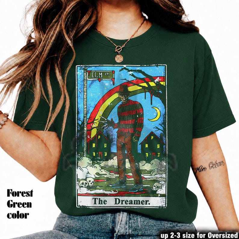 May include: Forest green t-shirt with a tarot card design. The graphic features the text "ELM ST" and "The Dreamer," along with a figure in a striped sweater, a rainbow, and a crescent moon. Available in up to 2-3 sizes for an oversized fit.
