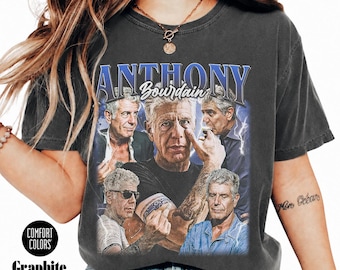 May include: A charcoal grey short-sleeve t-shirt featuring a graphic print of Anthony Bourdain in multiple poses. The text "ANTHONY" appears in large blue letters above his name in white script. Lightning bolt accents surround the images.