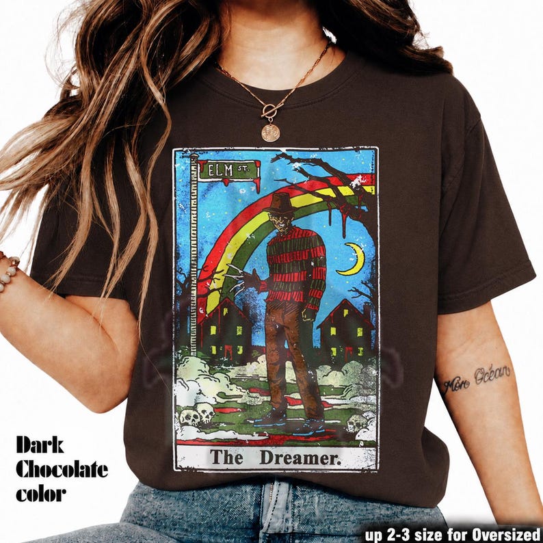 May include: Dark chocolate-coloured t-shirt with a tarot card-style graphic. The graphic shows a figure with a hat and striped sweater, a rainbow, and the text "ELM ST" and "The Dreamer." The shirt is available in up 2-3 sizes for an oversized fit.