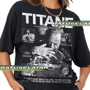 May include: Black t-shirt with the film title "TITANE" and a poster design. The design includes images of actors Vincent Lindon and Agathe Rousselle, and the text "A Film by Julia Ducournau".
