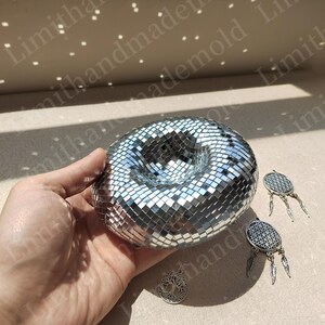 May include: A silver disco ball shaped like a donut with a hole in the center. The disco ball is made of small, mirrored tiles. There are three silver charms with a tree of life, a dreamcatcher, and a flower of life design.