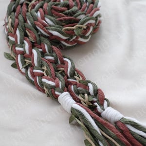 May include: Braided belt in shades of green, white, and burgundy. The belt is made of woven rope and features decorative leaf accents. The ends of the belt are finished with white tassels. A decorative accessory.