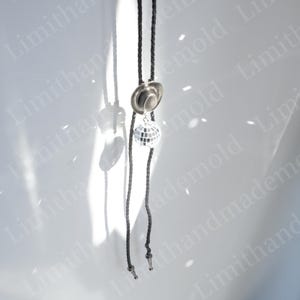 May include: A black bolo tie with a silver hat and disco ball charm. The tie is hanging against a white background.