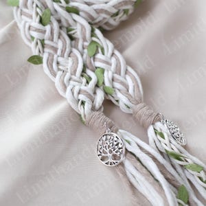 May include: Braided hair accessory featuring white, tan, and green accents. The braids are adorned with small green leaves and silver tree of life charms. The accessory is designed for hair styling.