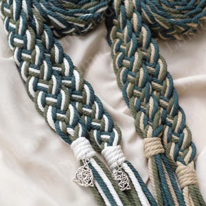Peut inclure: Ceinture en corde tressée dans les tons de vert, blanc et beige. La ceinture est ornée d'un charm en forme de nœud celtique à chaque extrémité. La corde est tissée de manière complexe, créant un accessoire texturé et décoratif.