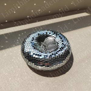 May include: A silver mirrored disco ball shaped trinket dish with a silver geometric design pendant resting inside.