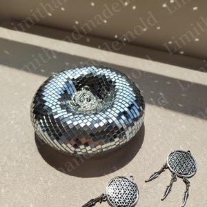 May include: A silver mirrored mosaic dish with a tree of life pendant inside. Two silver dreamcatchers with a flower of life design are on a beige surface.