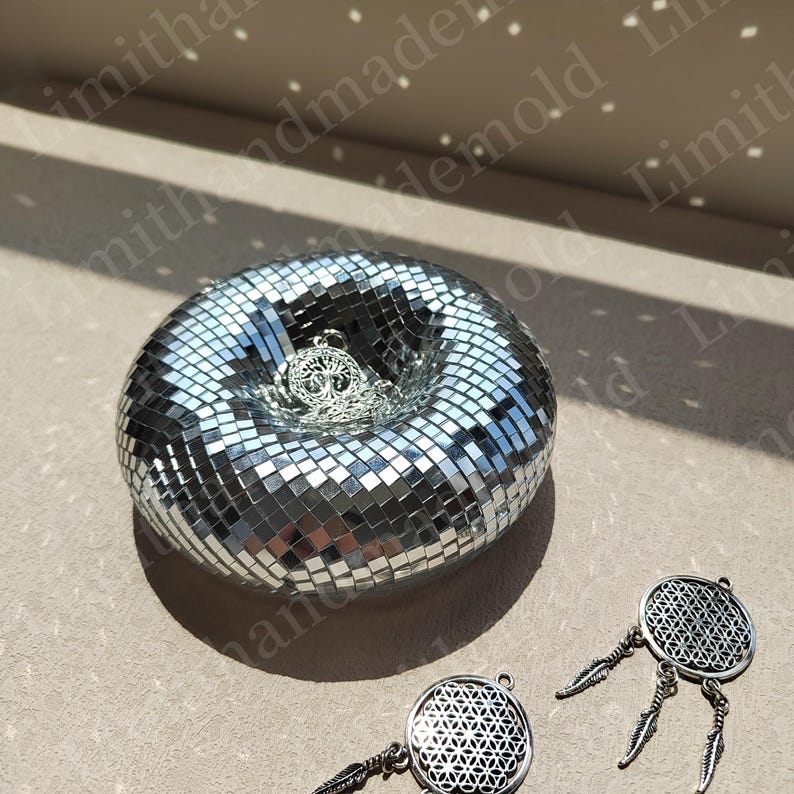 May include: A silver mirrored mosaic dish with a tree of life pendant inside. Two silver dreamcatchers with a flower of life design are on a beige surface.