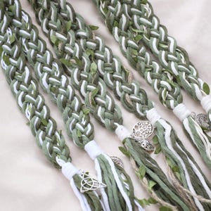 May include: Braided hair accessories in shades of green and white, adorned with small leaf accents and silver charms. The braids are finished with white tassels and decorative metal pendants, creating a natural, bohemian look.