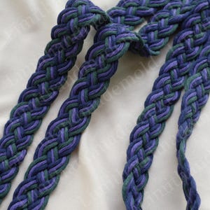 May include: Close-up of a braided belt in shades of blue and green. The belt is made of thick, woven material, with a detailed pattern. The colors are interwoven, creating a textured appearance. The belt is likely for fashion or craft purposes.