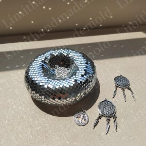 May include: A silver mirrored mosaic bowl with a silver metal charm inside. Three silver metal charms with feathers hang on the right side of the bowl.