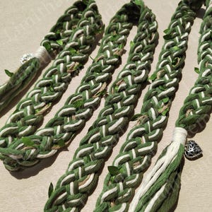 May include: A braided rope with green and white strands, decorated with small green leaves. The rope has a tassel at one end with a silver charm in the shape of a tree.