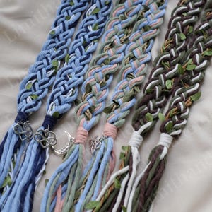 May include: Braided hair extensions in various colors, including blue, pink, green, and brown. Each braid features decorative charms and small green leaf accents. The braids are arranged side by side, showcasing their intricate designs.