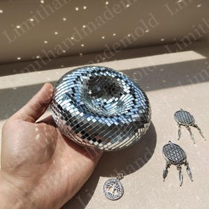May include: A silver disco ball with a hole in the center, held in a hand. Three silver charms with feather accents are also pictured.