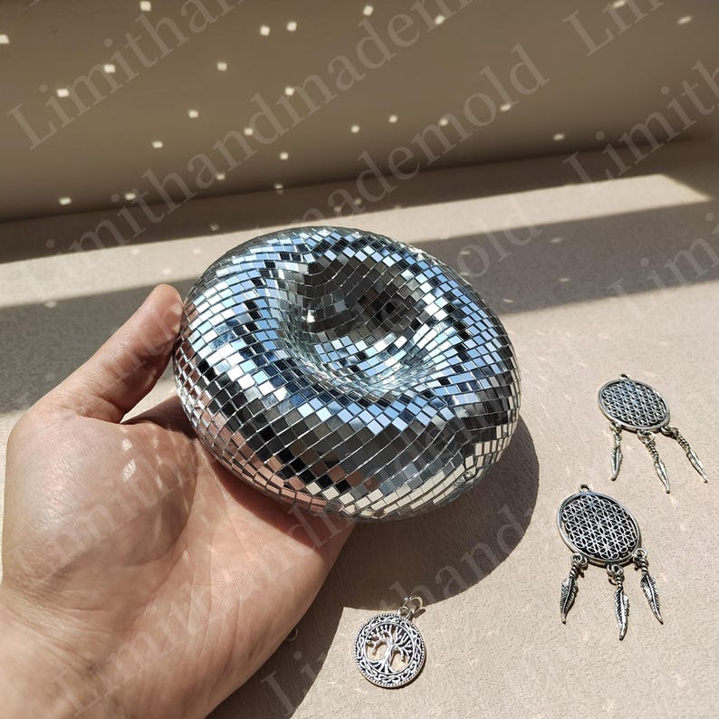 May include: A silver disco ball with a hole in the center, held in a hand. Three silver charms with feather accents are also pictured.