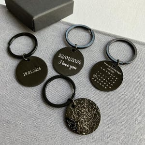 Könnte beinhalten: Eine Sammlung schwarzer Metall-Schlüsselanhänger mit runden Anhängern. Jeder Schlüsselanhänger hat ein anderes Design, darunter Daten, der Satz "I love you", ein Kalender und eine Sternenkarte. Die Schlüsselanhänger haben schwarze Ringe und werden auf einer grauen Oberfläche präsentiert.