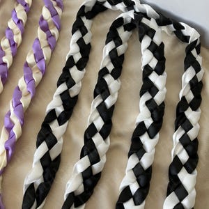 May include: Braided satin ribbons in black and white, and purple and cream. The ribbons are woven together in a thick, flat braid. The black and white ribbons are in the foreground, while the purple and cream ribbons are in the background.