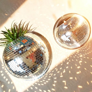 Puede incluir: Dos macetas de bola de discoteca, una con follaje verde. Las macetas son redondas con una superficie de mosaico espejado. La luz del sol proyecta un patrón de luces y sombras sobre la superficie.