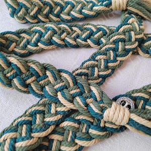 May include: A braided rope belt with a green and beige pattern. The belt has a metal tree of life buckle.