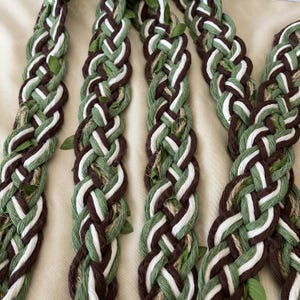 May include: Close-up of several braided strands. Each strand is a combination of brown, white, and green fibers, with small green leaves interspersed throughout. The braids are arranged in a parallel pattern, creating a textured and natural aesthetic.