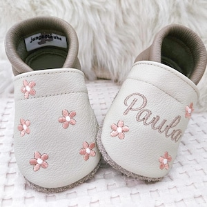 Könnte beinhalten: Ein Paar hellgraue Lederschuhe für Babys mit rosa gestickten Blumen und dem Namen "Paula" auf einem Schuh gestickt. Die Schuhe haben einen braunen Lederbesatz am oberen Rand.