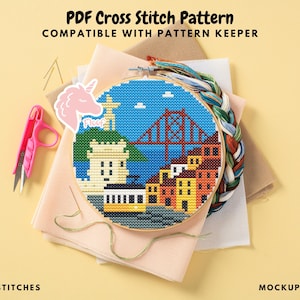 May include: A cross-stitch pattern in a wooden hoop depicts a cityscape with a bridge, buildings, and a tram. The pattern is surrounded by embroidery floss, fabric, pink scissors, and a unicorn sticker. Text on the image reads "PDF Cross Stitch Pattern".